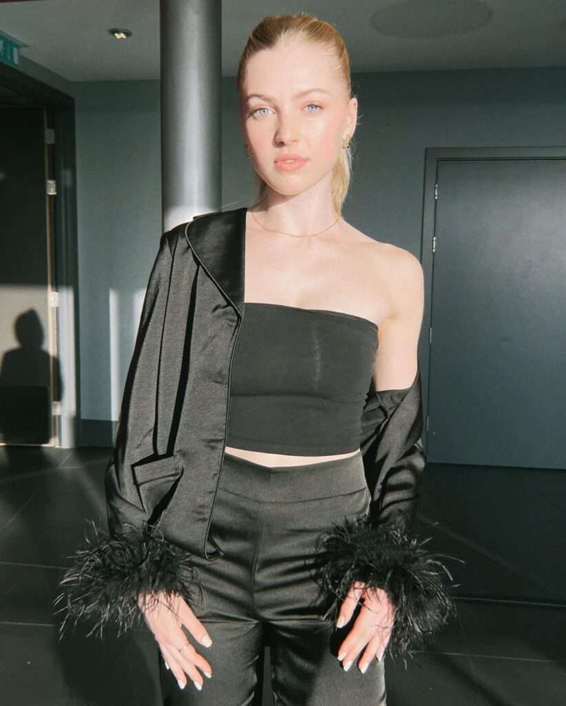 Annie Knowles is wearing a black off shoulder crop top with matching silk jacket and pants while looking towards camera 