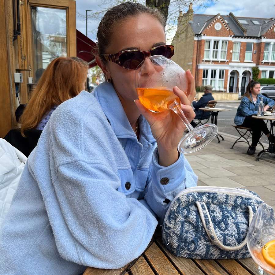 Jena Goldsack is drinking juice while wearing a sky blue woollen long coat 