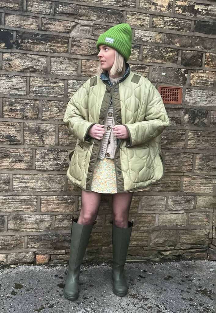Sarah Hughes in a reformation style dress while wearing a sweater, over coat pair with a green cap and boots 