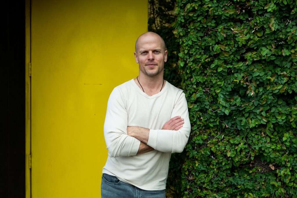 Tim Ferriss taking a picture with a beautiful smile and wearing a white full sleeves t-shirt

