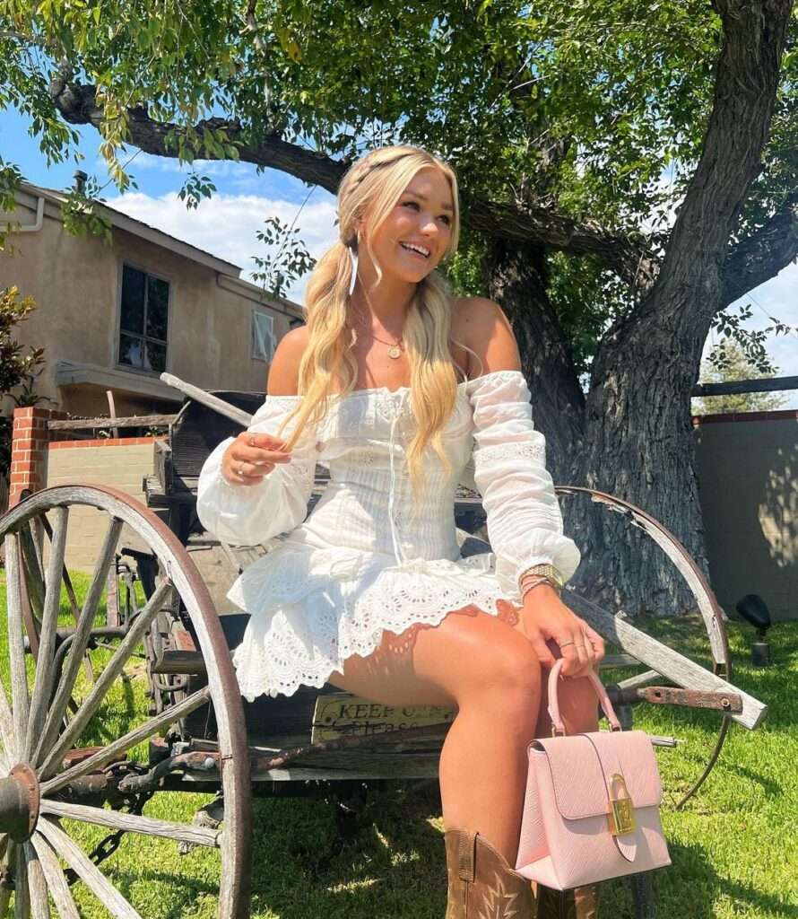 Katie sigmond in a white chic reformation style dress pair with pink bag and brown boots while poses for a picture