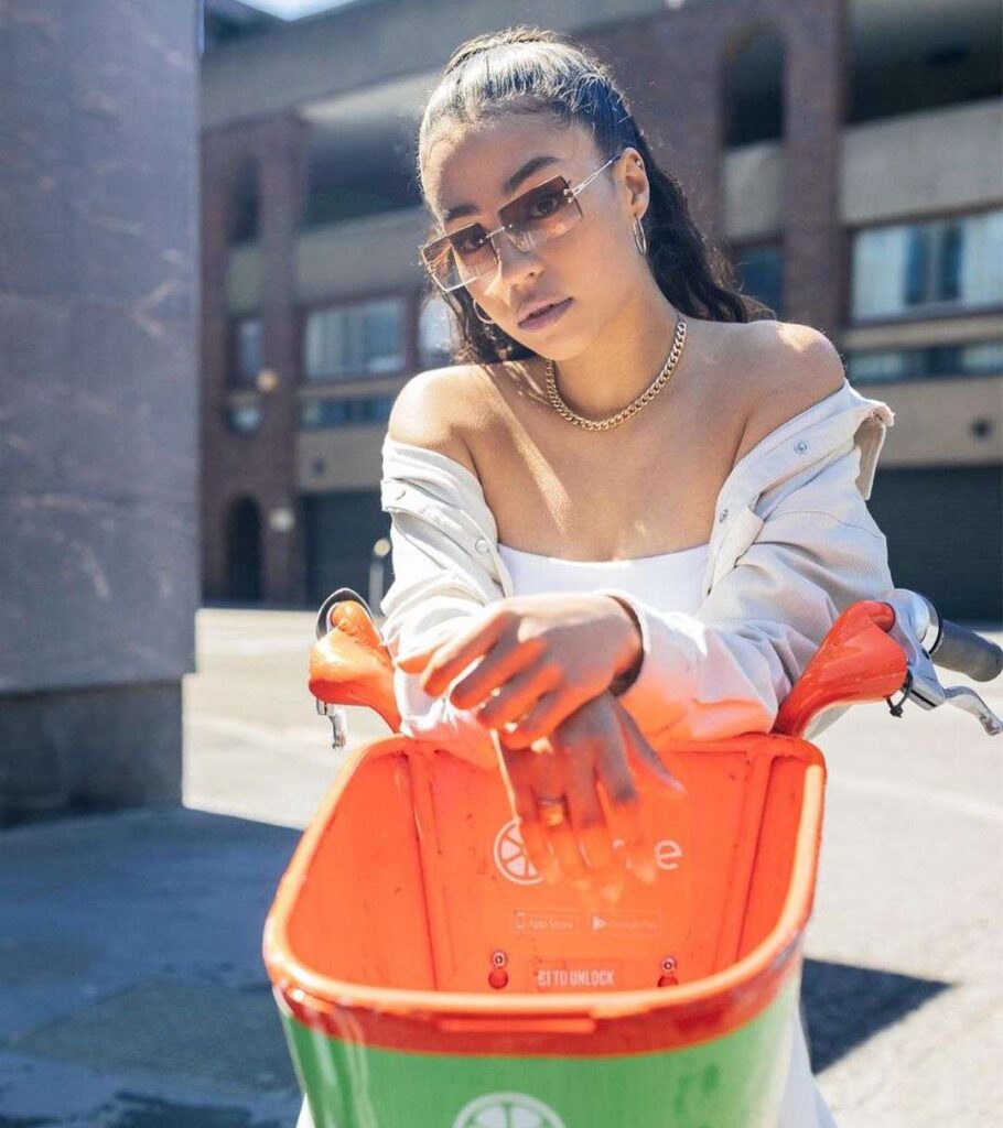 Lirian Santos in a off white top pair with a chain necklace, small hoops, and goggles while sitting on a bike