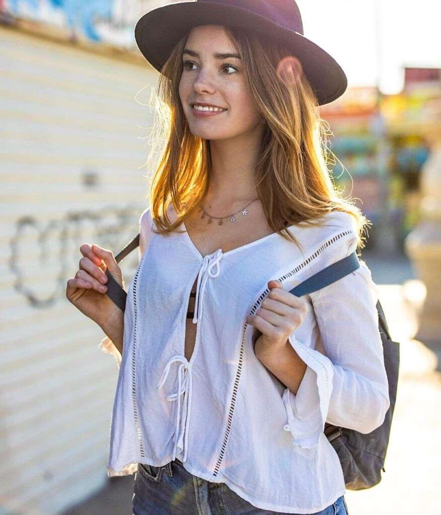 Makenzie Raine in the white top pair with denim shorts and maroon hat while smiling towards camera
