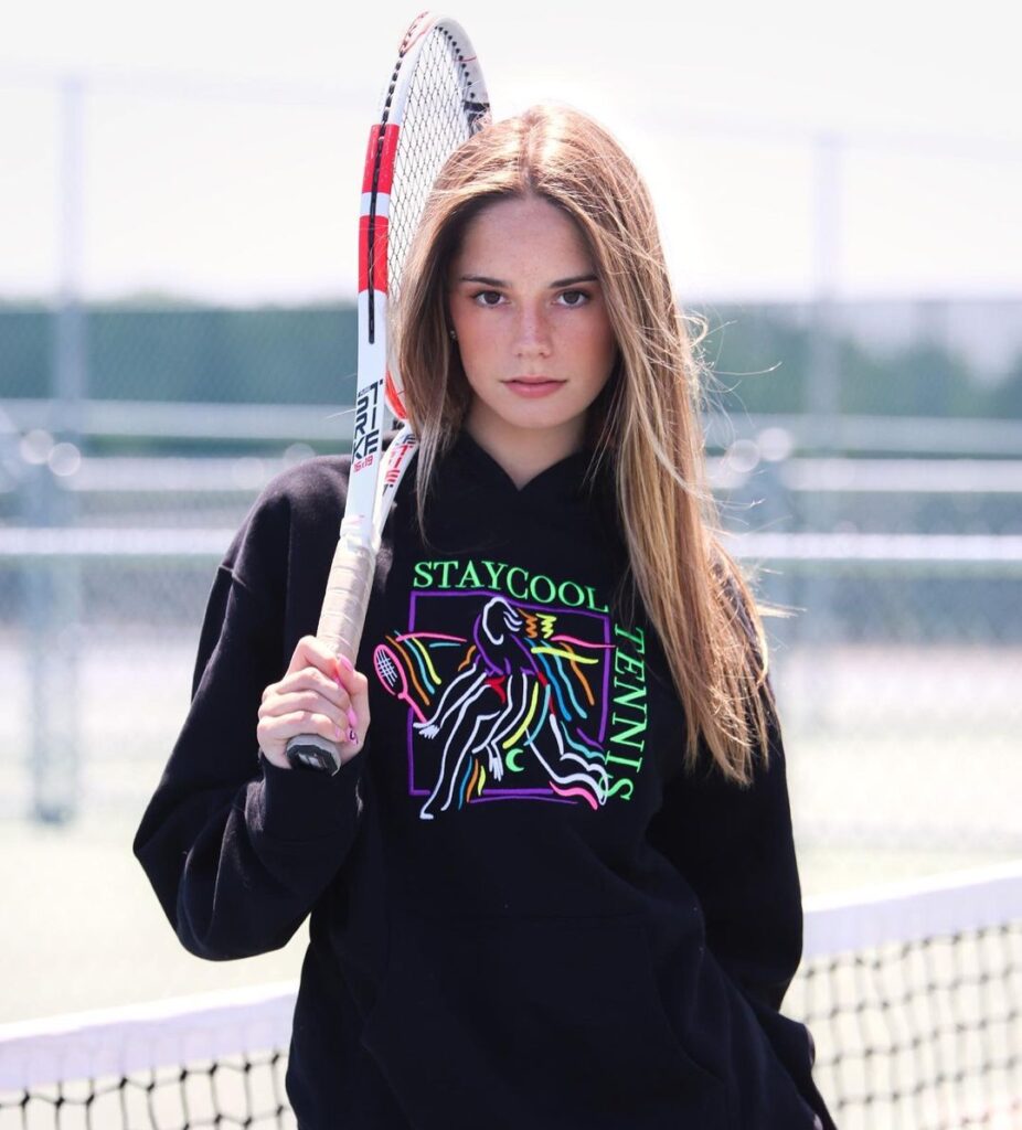 Makenzie Raine in a black hoodie pair with red trouser while holding a racket in her hand and looking towards camera