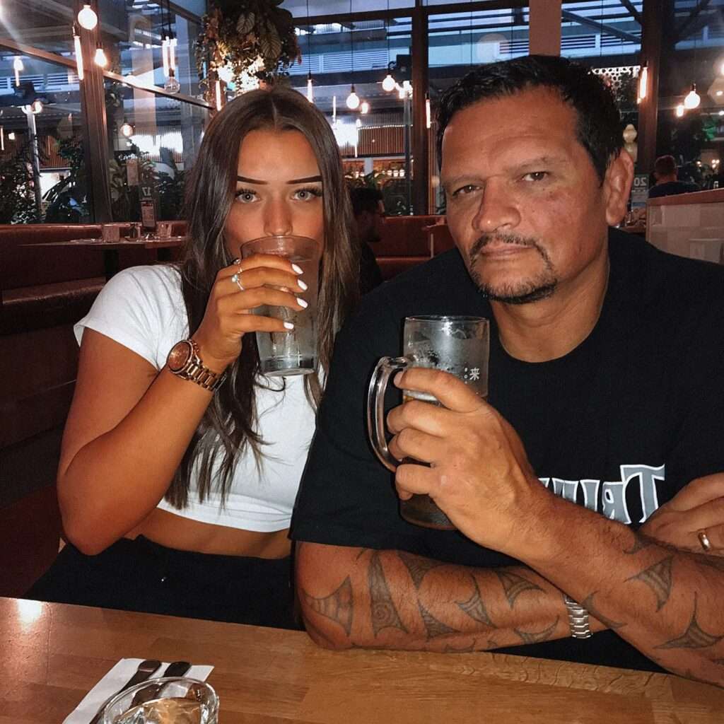 Tui jones in a white crop t-shirt with black jeans holding a glass in her hand while taking selfie with her father