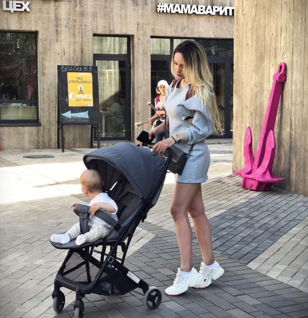 Yana Vasileva in a grey jumpsuit pair with white joggers while holding her baby stroller