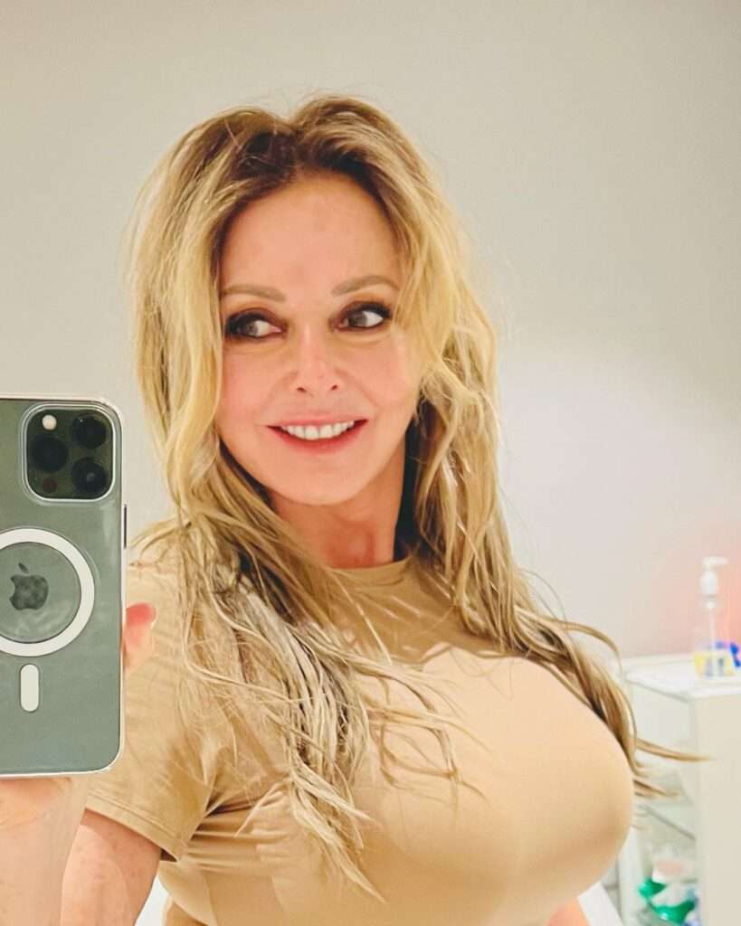 Carol Vorderman in brown t-shirt while taking a selfie in the front of mirror