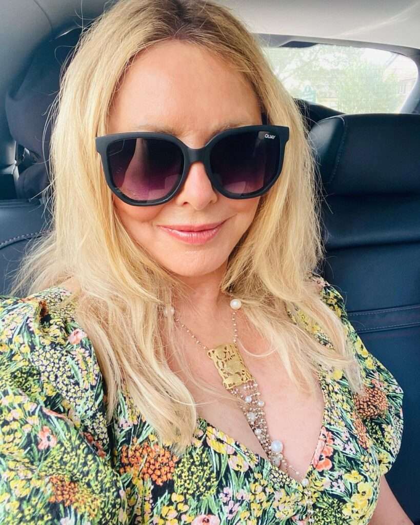 Carol Vorderman poses for a picture while sitting in her car and wearing a printed top with deep neckline
