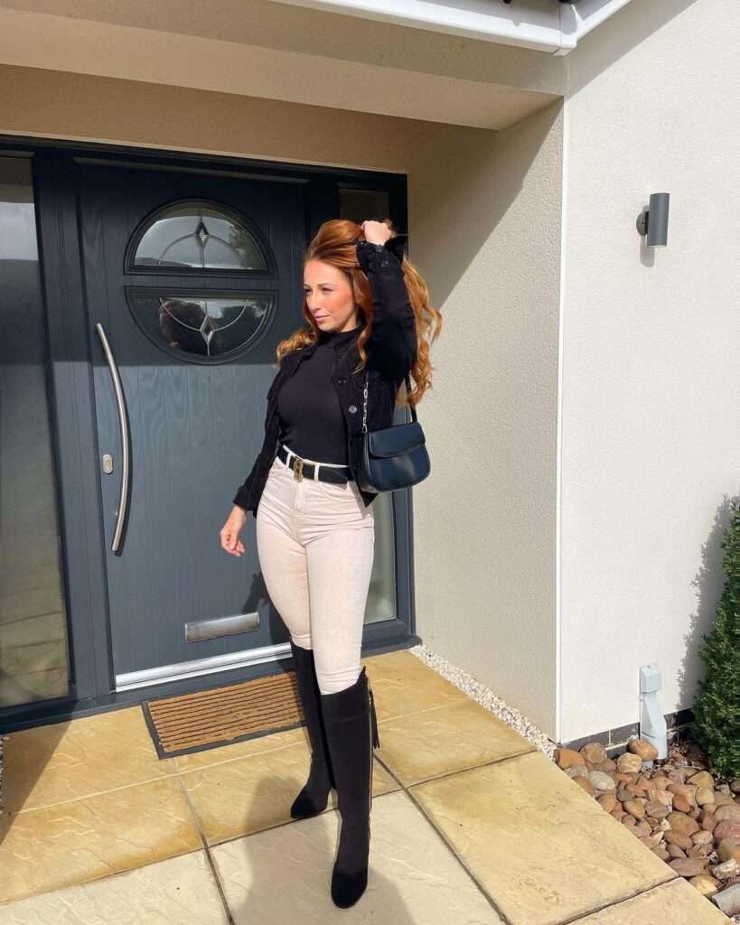 Melissa Carroll wearing a black high neck shirt with creamy brown pants, long boots, and black bag, while poses for a photo