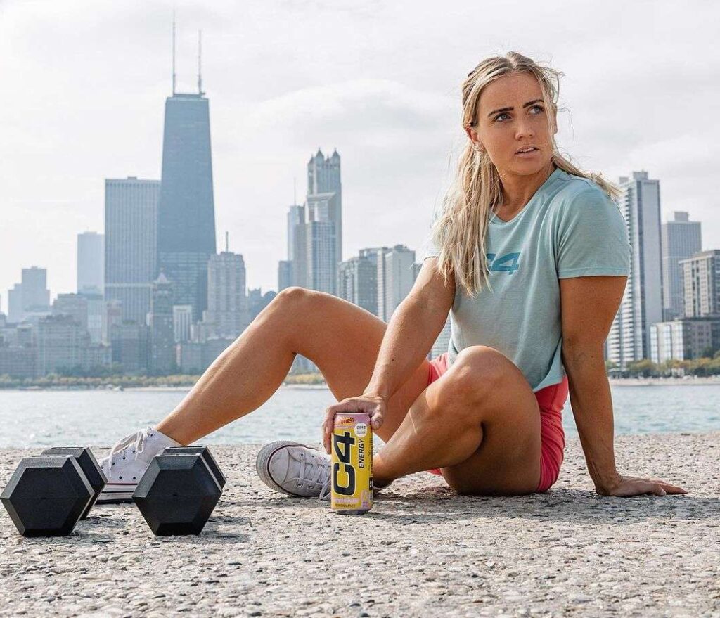 Sarah Hryniewicz is sitting on a road as she is posing for a picture while showing her collaboration for C4 Energy while wearing a t-shirt with shorts.