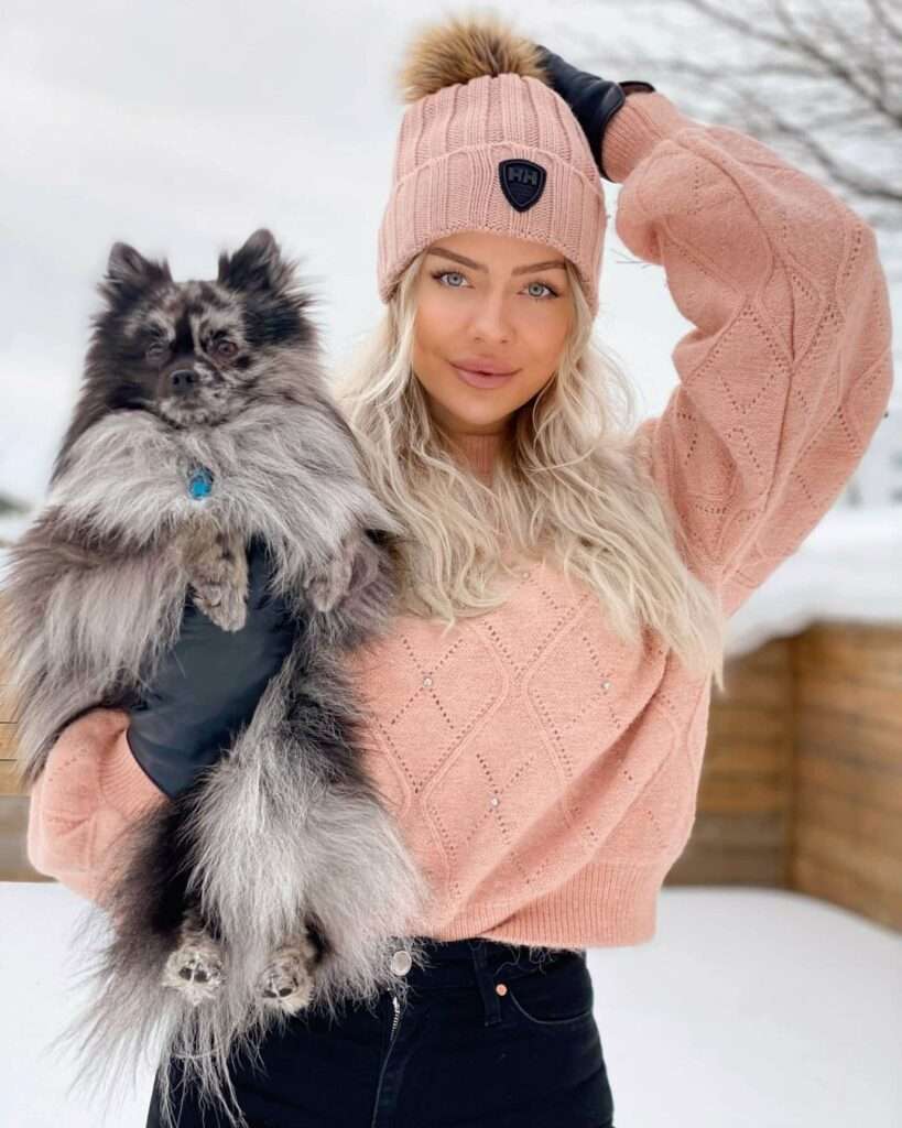 Trine Petronella is posing for a picture and she is standing in a snow with her dog while wearing a jersey and cap with jeans.