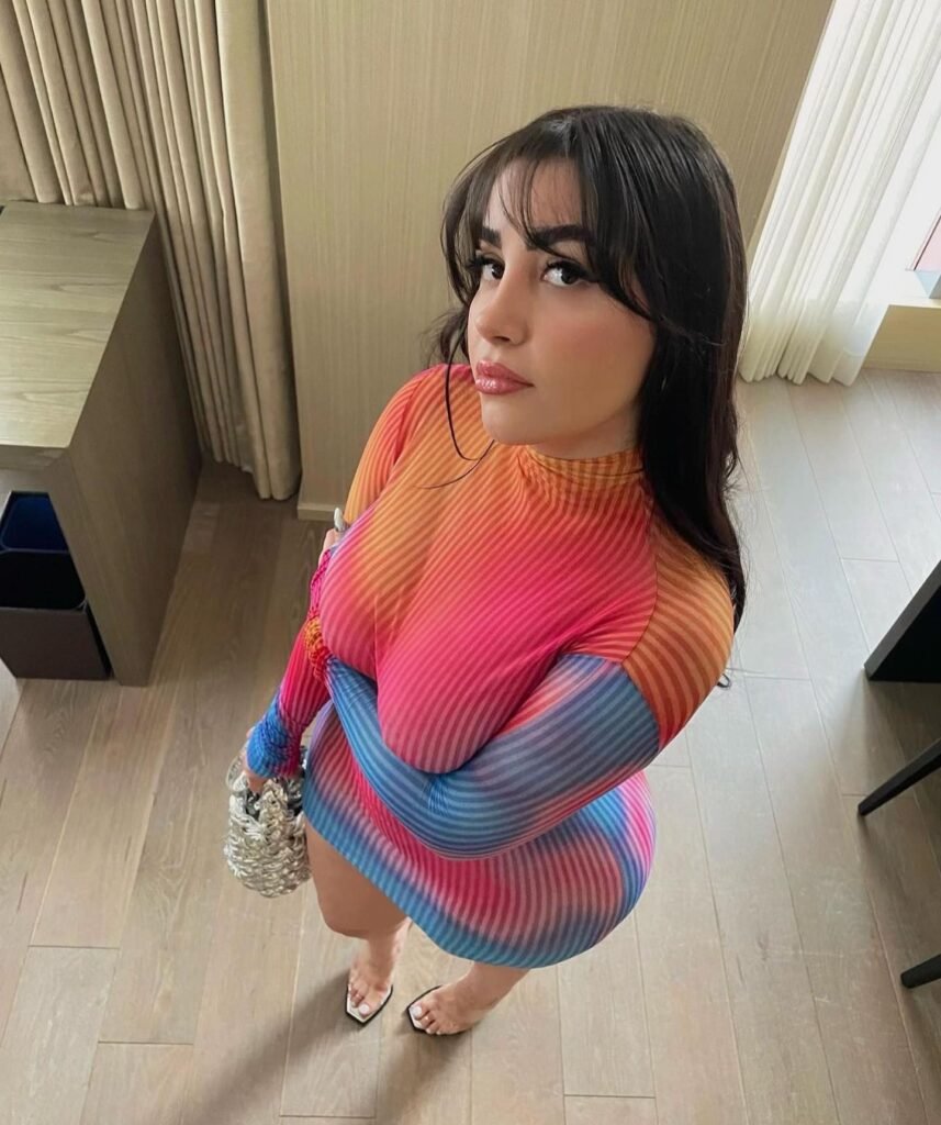 Who is Amiliasoforiegn? Bio, Wiki, Age, Height, Net worth, Onlyfans, 23 Amelia Curvy posing indoors in a vibrant, form-fitting striped dress with pink, orange, and blue tones, holding a silver handbag.