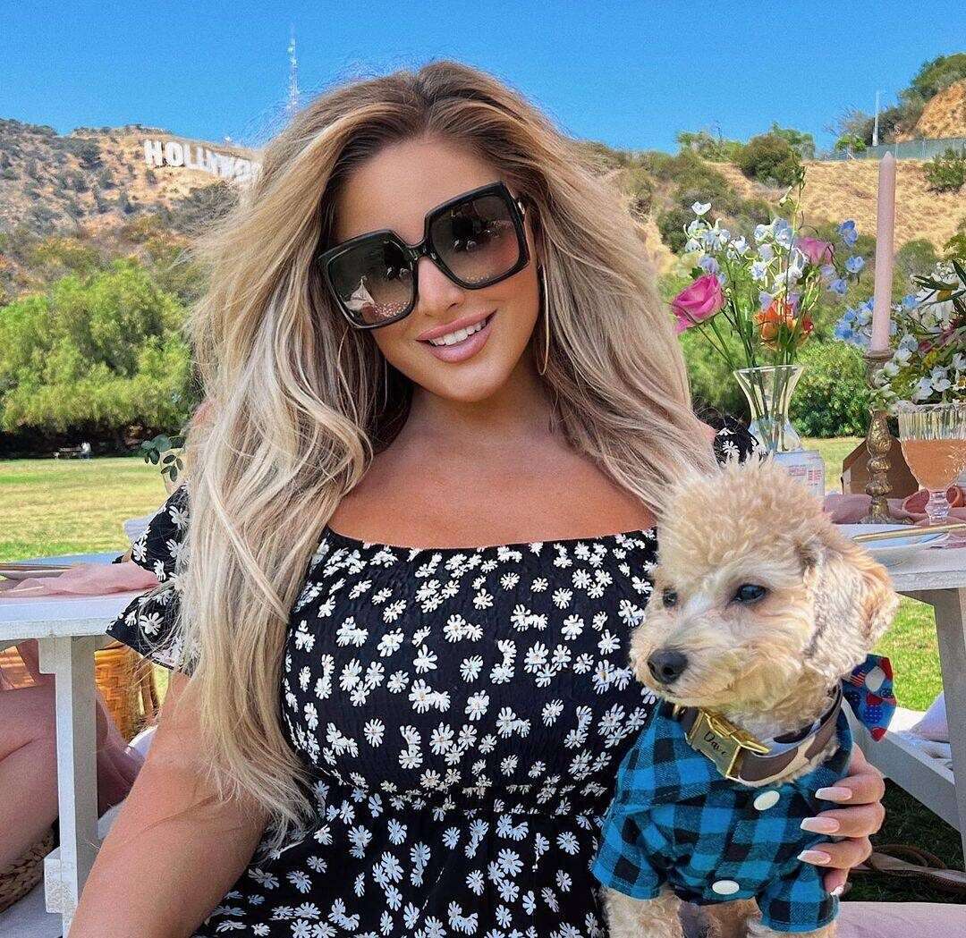 Ashley Alexiss in a black floral top while holding her pup and smiling towards camera