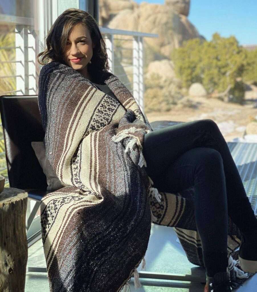 Colleen Ballinger is sitting on a chair and here she is wearing a shirt and pant while wrapping a shawl and posing for a picture.