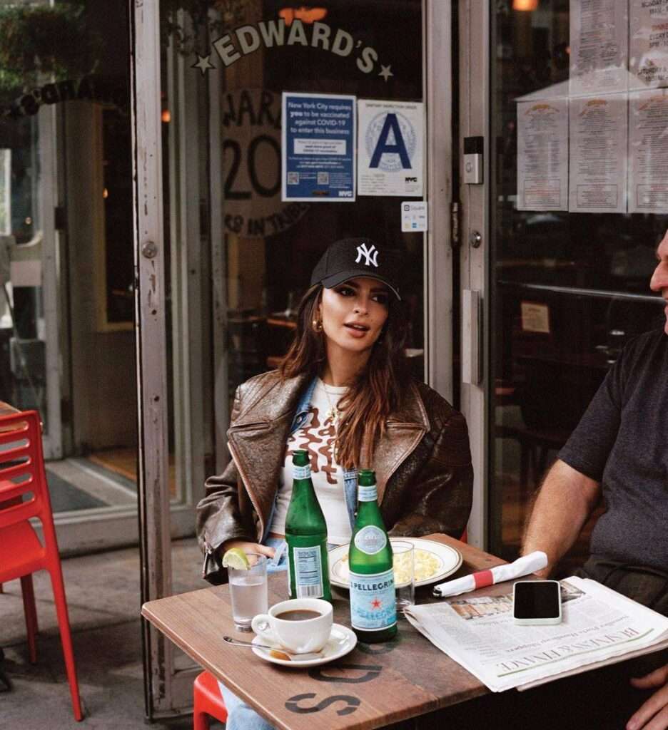 Emily Ratajkowski is wearing the white printed t-shirt with leather coat and cap while discussion in the meeting