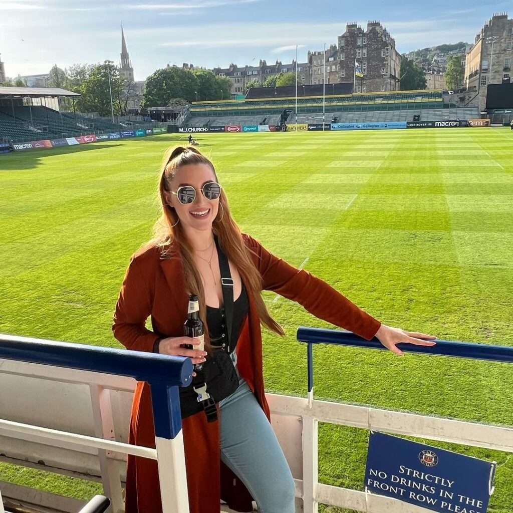 Emily Stallabrass in the black tank top pair with blue jeans and brown log coat while standing on the stadium