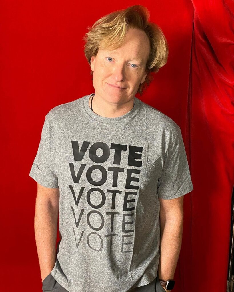 Conan O’Brien is looking smart and wearing the t shirt in front of red backgrounf posing for the picture