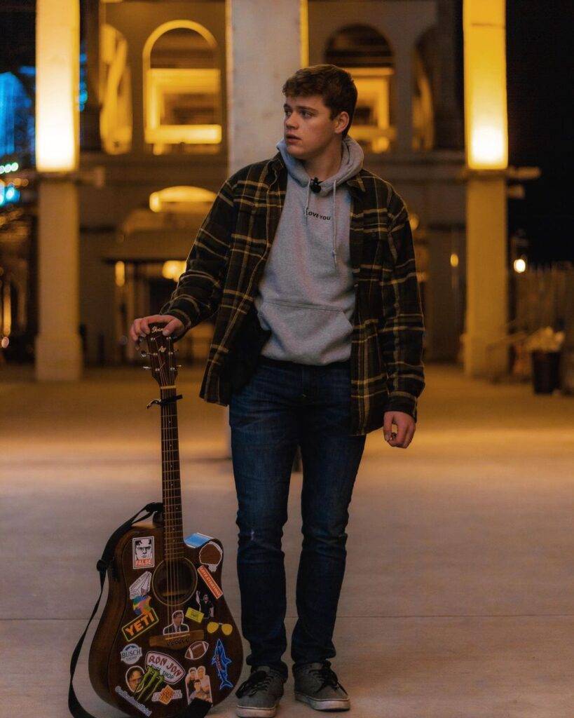 Will Cullen is wearing grey hoodie over jacket and holding the guitar and posing for the picture