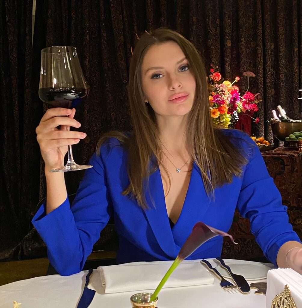 Svetlana in the blue coat pair with black inner while holding a glass of wine in her hand and looking towards camera