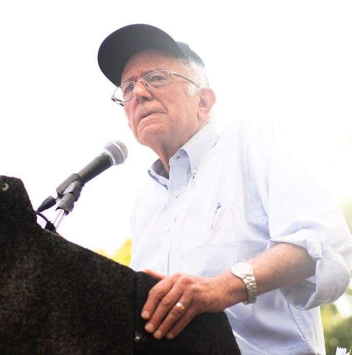 Bernie Sanders is standing and is addressing with his nation on issues and is wearing a checkered shirt.
