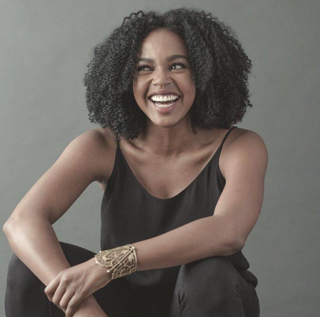 Jerrika Hinton is smiling while wearing a black Binyan shirt with pant.