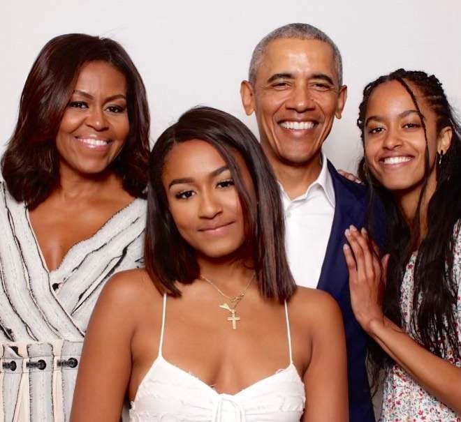 Michelle Obama is smiling and is posing for a picture as she is standing with her family including her two daughter and husband
