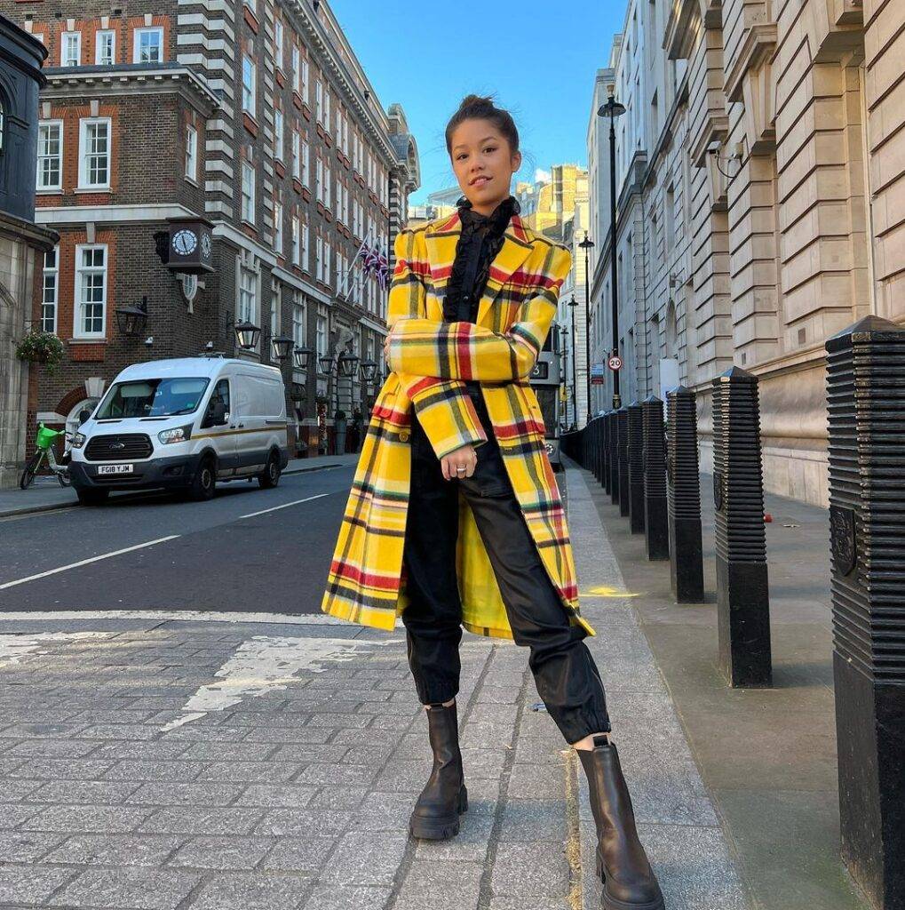 Trinity Jo-Li Bliss in the black outfit pair with yellow coat and black shows while looking towards camera