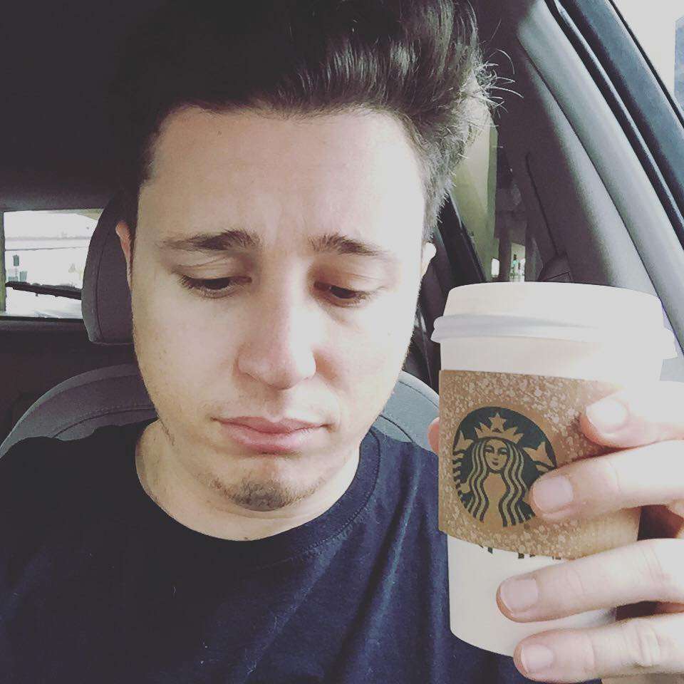 Corey B is taking coffee and is sitting in his car and is posing funny for a picture.