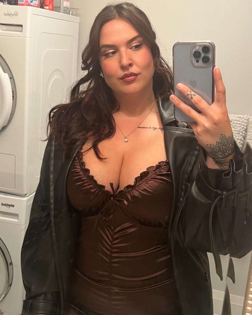 Steph Oshiri is wearing brown dress over black jacket and standing in front of mirror while taking selfie
