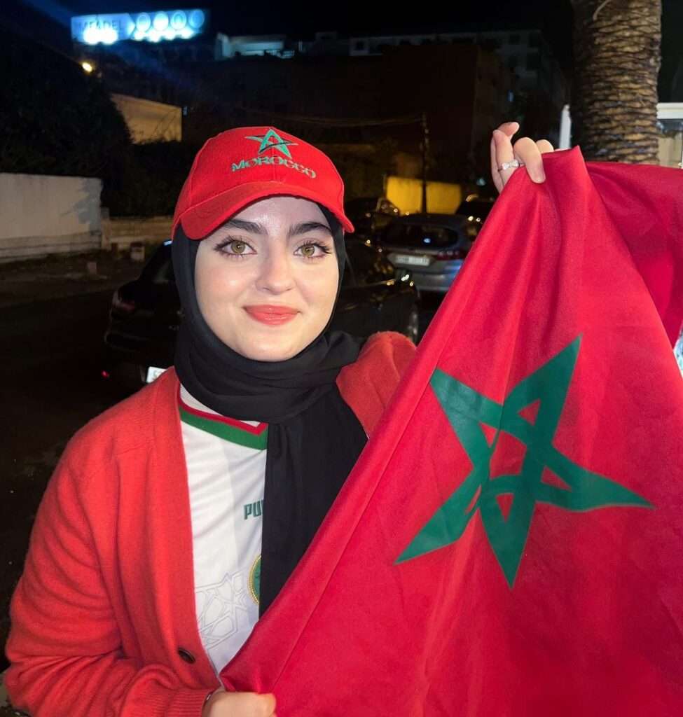 Amina Dehbi in the white t-shirt pair with red sweater and red cap while showing Morocco fag towards camera