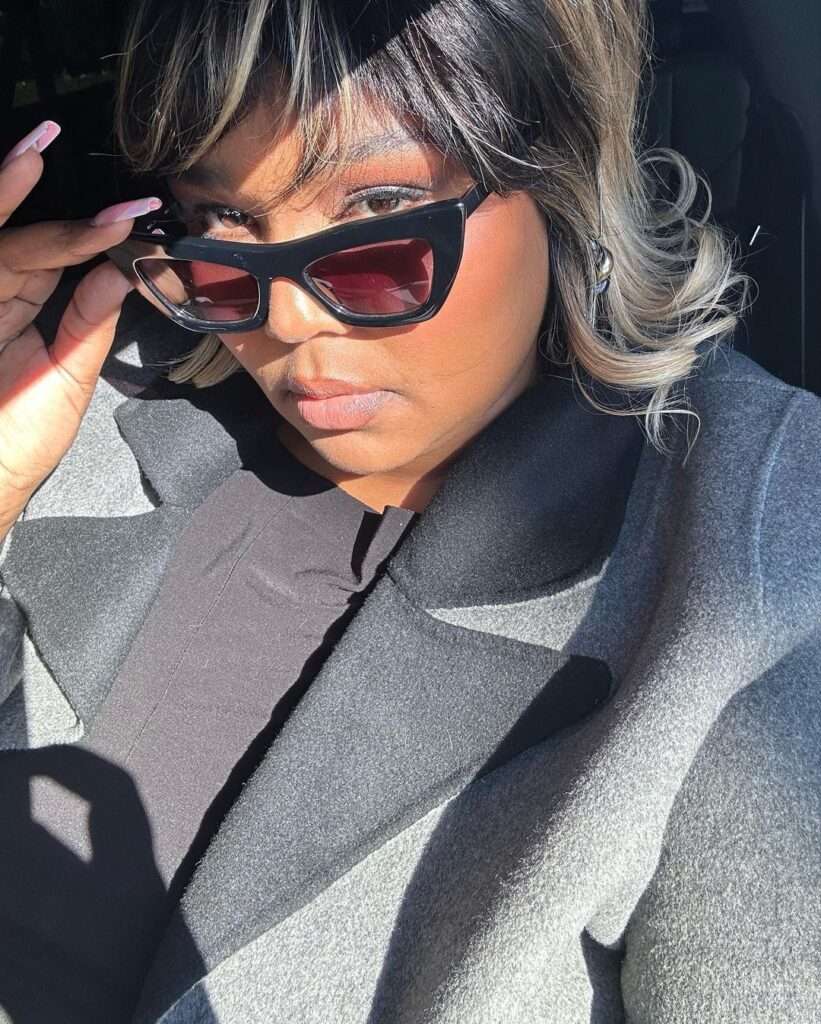 Lizzo is wearing black coat and glasses or sitting in the car while posing for taking picture