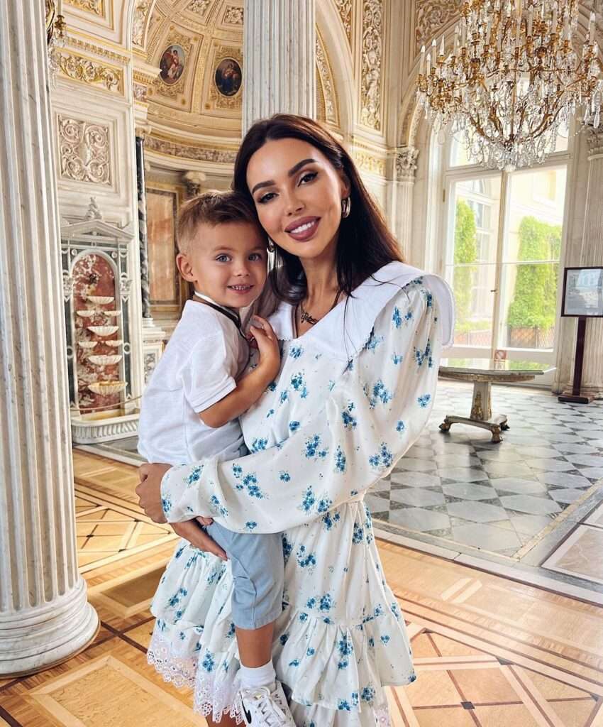 Oxana Samoylova is wearing white dress or holding his baby or smiling while posing for the picture