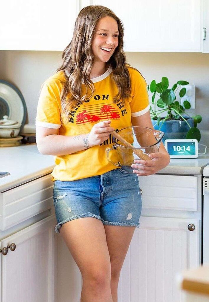 Bella Bucchiotti is standing in a kitchen and is telling a new recipe while wearing a shirt with shorts.