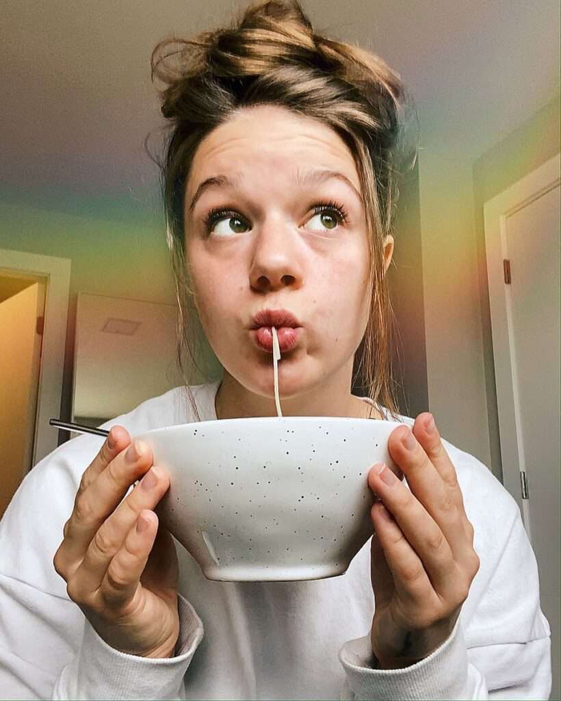 Bella Bucchiotti is eating noodles and is posing for a picture while wearing a t-shirt.