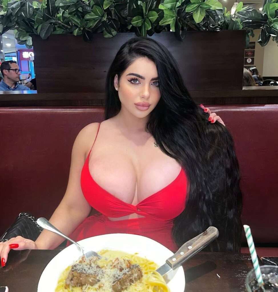 Adriana Alencar is wearing red dress and sitting in front of tasty food or posing for taking the picture