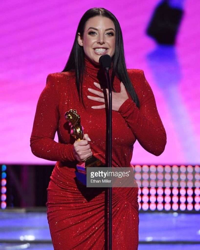 Angela White is wearing red dress and holding the trophee or standing in front of mike or posing for the picture