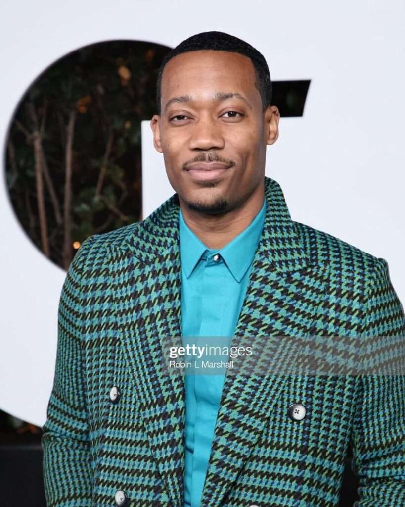 Tyler James WIlliams is wearing blue shirt over coat or smiling while posing for the picture