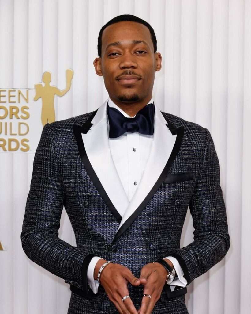 Tyler James Williams is wearing white shirt over pant and coat or smiling while posing for the picture