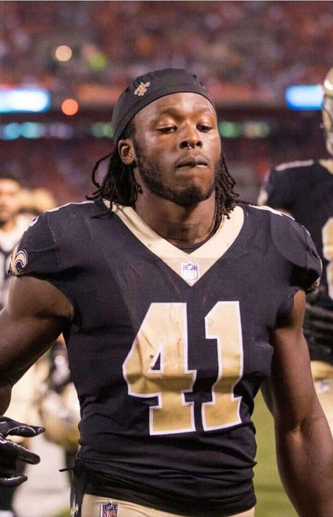 Alvin Kamara is wearing black t shirt and standing in the ground or posing while taking the picture