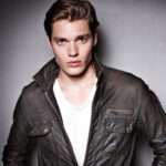 Dominic Sherwood is wearing white shirt over black jacket or posing while taking the picture