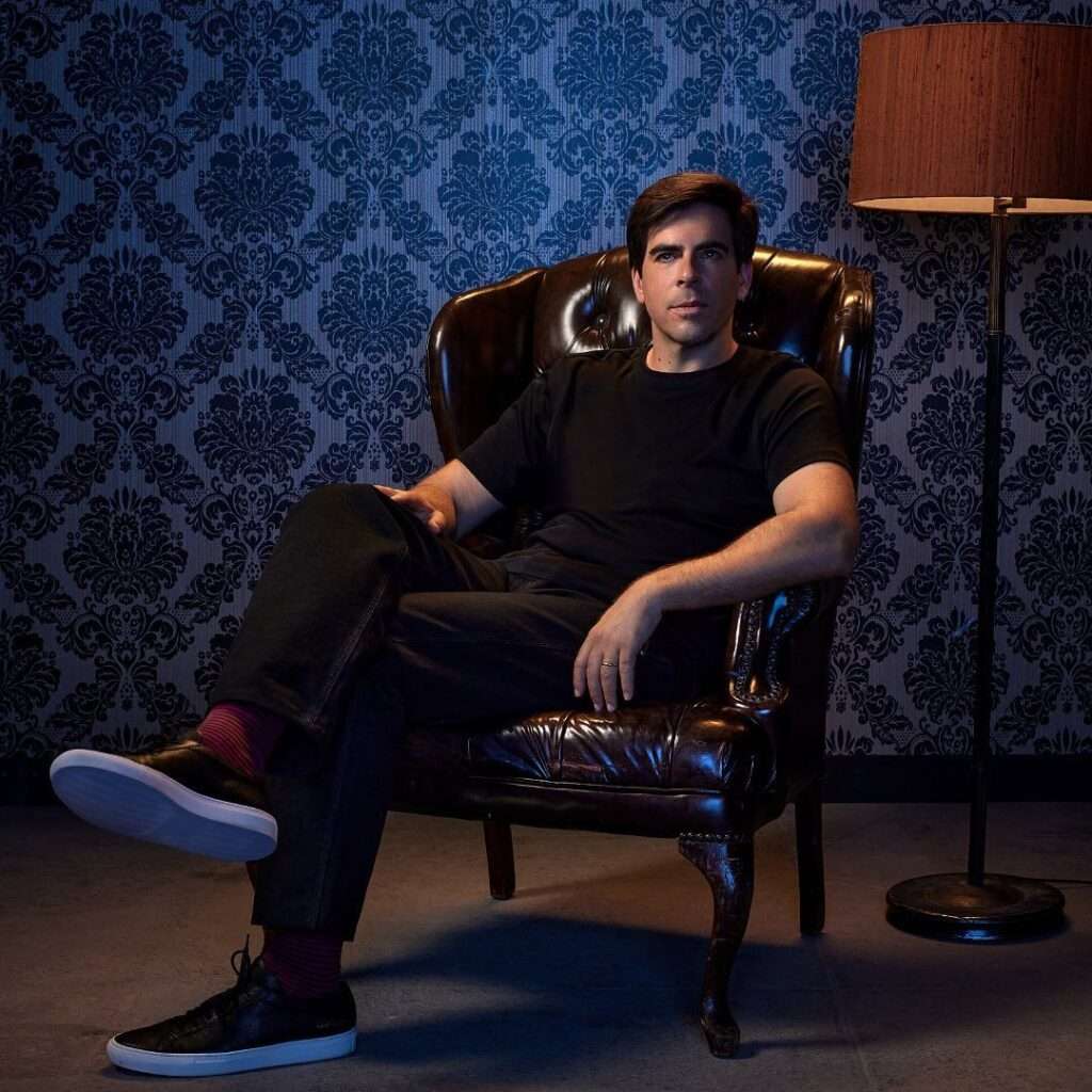Eli Roth is wearing black t shirt and trouser or sitting on the chair and posing for the picture