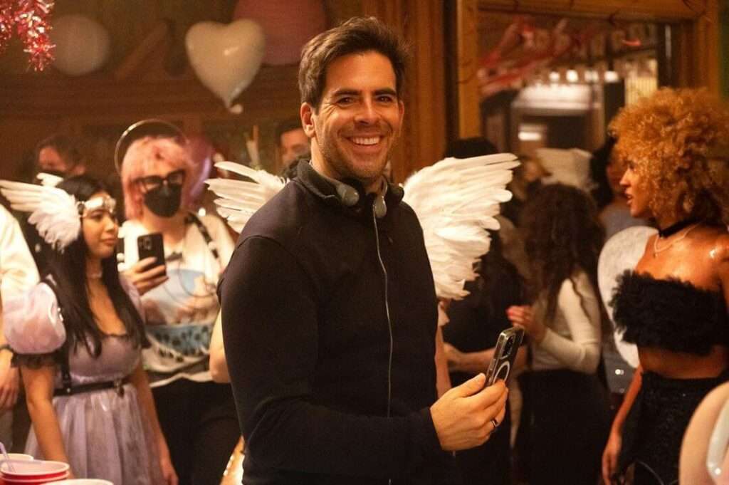 Eli Roth is wearing black outfit and holding the phone and smiling while posing for the picture