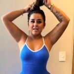 Close-up of Dinalva Vasconcelos tying her hair in a blue ribbed tank top, showing her tattoo and wearing gold jewelry indoors