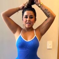 Close-up of Dinalva Vasconcelos tying her hair in a blue ribbed tank top, showing her tattoo and wearing gold jewelry indoors