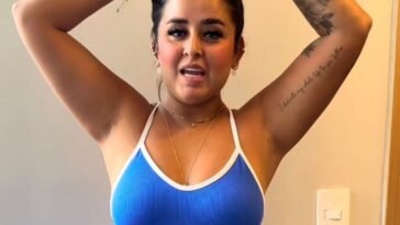 Close-up of Dinalva Vasconcelos tying her hair in a blue ribbed tank top, showing her tattoo and wearing gold jewelry indoors