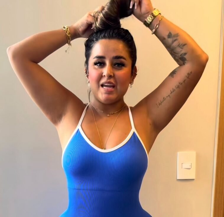 Close-up of Dinalva Vasconcelos tying her hair in a blue ribbed tank top, showing her tattoo and wearing gold jewelry indoors