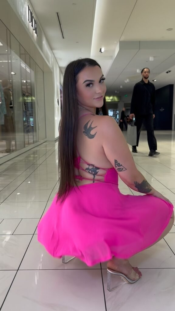 Kiley Lossen crouching playfully in a bright pink strappy dress and clear heels inside a luxury shopping mall, revealing tattoos and confident energy