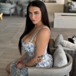 Kiley Lossen sitting elegantly in a floral summer dress, showcasing her natural beauty and tattoos while posing on a patio chair.
