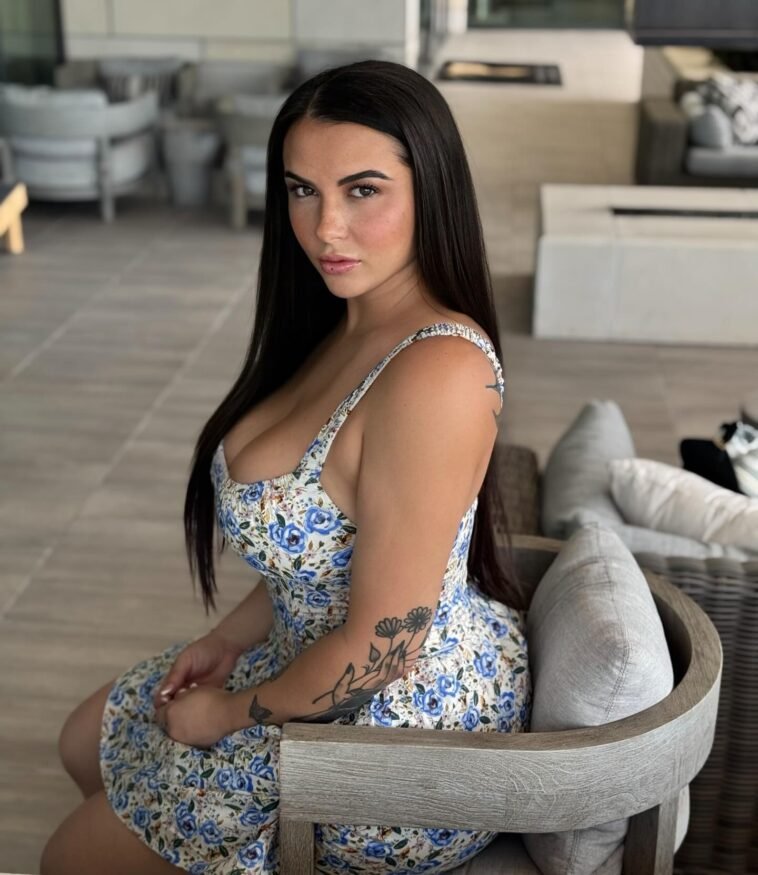 Kiley Lossen sitting elegantly in a floral summer dress, showcasing her natural beauty and tattoos while posing on a patio chair.