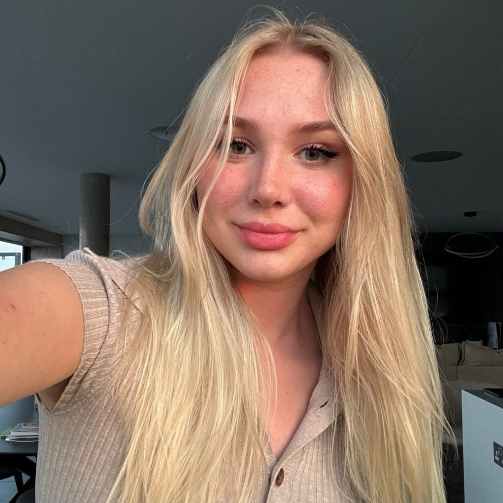 Patty Peach, a young woman with long, straight blonde hair and natural makeup, smiles slightly while taking a selfie indoors. She’s wearing a beige ribbed top with buttons, and soft lighting highlights her fresh, glowing skin.